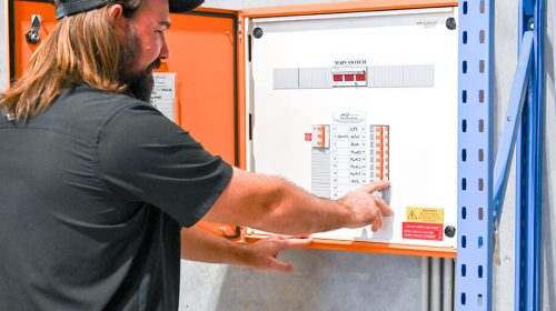 Residential electrician Loganholme technician testing a living room power point with a digital meter while a family watches nearby.